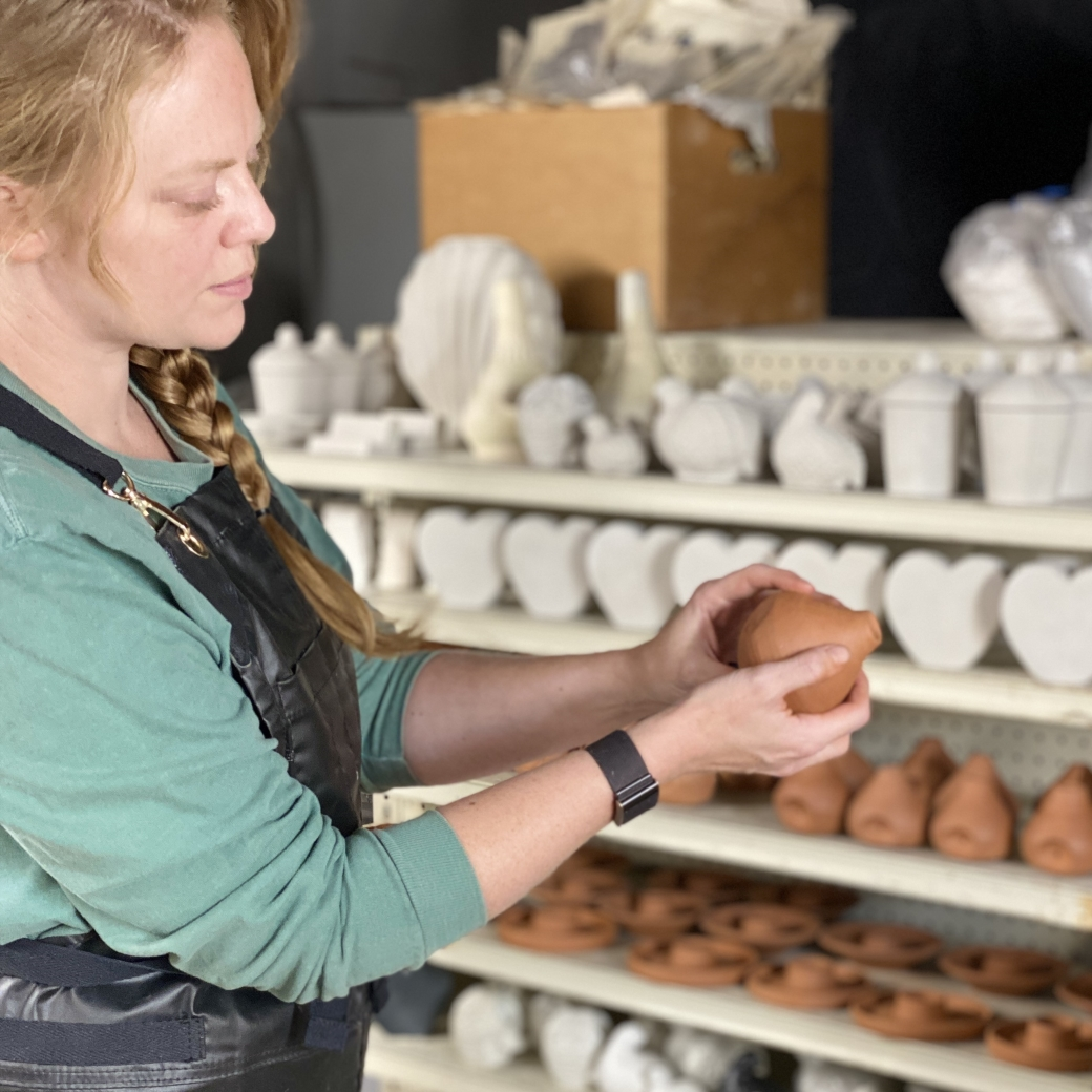 Female holding a piece of pottery standing in a studio in front of a shelf of in progress work.